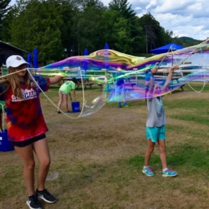 Participants making large iridescent bubbles with BubbleStix wooden wand sets on a grassy activity field