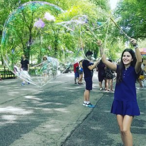 Woman in a navy dress using a wooden BubbleStix wand to make a giant bubble in a leafy park